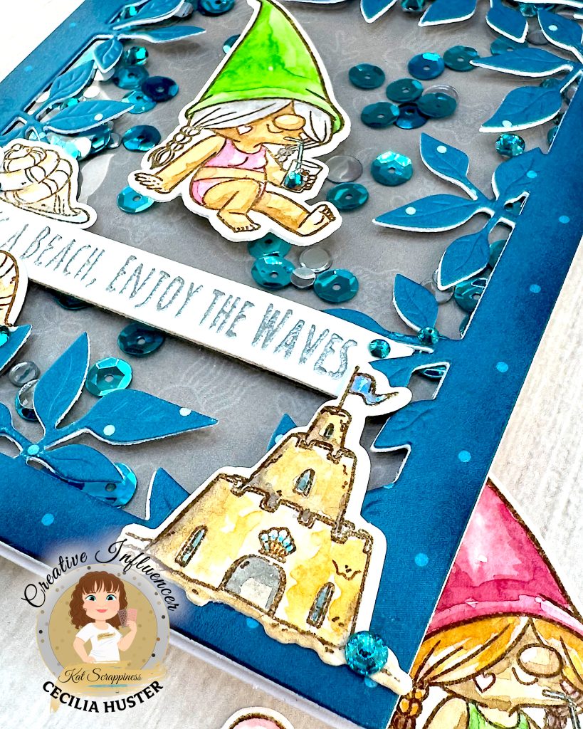 A handmade greeting card with a beach gnome theme displayed against a pale wooden surface. The card features a clear window filled with turquoise and silver sequins. The window is bordered by a teal, leafy frame.
The main design shows a cute gnome with long, grey hair and a bright green pointed hat. She appears to be relaxing in a bikini while holding a small drink. A banner across the card reads "LIFE'S A BEACH, ENJOY THE WAVES!" The banner is decorated with sea shells. A sandcastle has been placed in the lower right corner.
The photo is taken from the lower right corner of the card. Under the card another gnome figure with a pink hat can be glimpsed.
The bottom right corner shows the "Kat Scrappiness Creative Influencer CECILIA HUSTER" logo featuring a cartoon woman with brown hair.