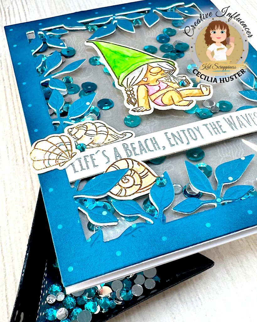A handmade greeting card with a beach gnome theme displayed against a pale wooden surface. The card features a clear window filled with turquoise and silver sequins. The window is bordered by a teal, leafy frame.
The main design shows a cute gnome with long, grey hair and a bright green pointed hat. She appears to be relaxing in a bikini while holding a small drink. A banner across the card reads "LIFE'S A BEACH, ENJOY THE WAVES!" The banner is decorated with sea shells. A sandcastle has been placed in the lower right corner.
The card is photographed from the lower left corner. Under it a black triangle tray holding turquoise gems is visible.
The bottom right corner shows the "Kat Scrappiness Creative Influencer CECILIA HUSTER" logo featuring a cartoon woman with brown hair.