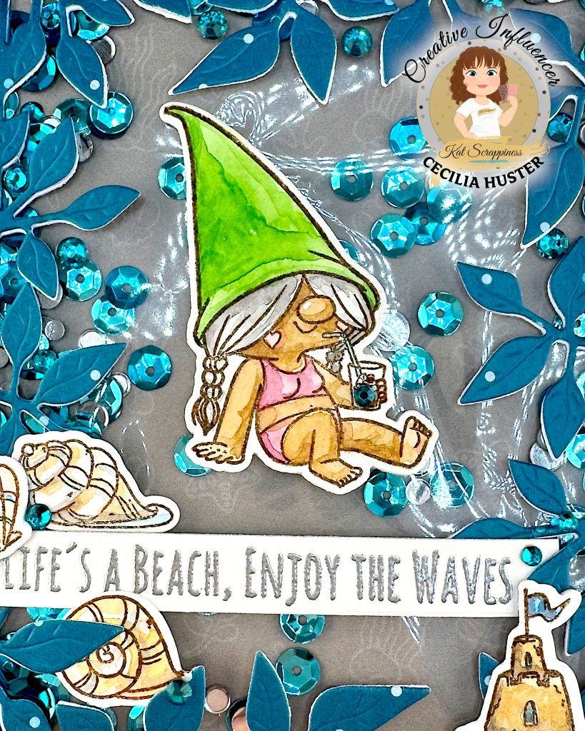 A handmade greeting card with a beach gnome theme displayed against a pale wooden surface. The card features a clear window filled with turquoise and silver sequins. The window is bordered by a teal, leafy frame.
The main design shows a cute gnome with long, grey hair and a bright green pointed hat. She appears to be relaxing in a bikini while holding a small drink. A banner across the card reads "LIFE'S A BEACH, ENJOY THE WAVES!" The banner is decorated with sea shells. A sandcastle has been placed in the lower right corner.
This is a closeup that allows us to see the detail of the watercolored gnome and the many turquoise sequins behind her.