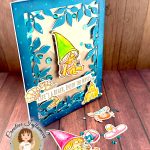 A handmade greeting card with a beach gnome theme displayed on a wooden surface. The card features a clear window filled with turquoise and silver sequins. The window is bordered by a teal, leafy frame. The main design shows a cute gnome with long, grey hair and a bright green pointed hat. She appears to be relaxing in a bikini while holding a small drink. A banner across the card reads "LIFE'S A BEACH, ENJOY THE WAVES!" The banner is decorated with sea shells. A sandcastle has been placed in the lower right corner. In front of the card are several coordinating die-cut elements including another gnome figure with a pink hat, a crab, and turquoise gems scattered around. The bottom left corner shows the "Kat Scrappiness Creative Influencer CECILIA HUSTER" logo featuring a cartoon woman with brown hair.