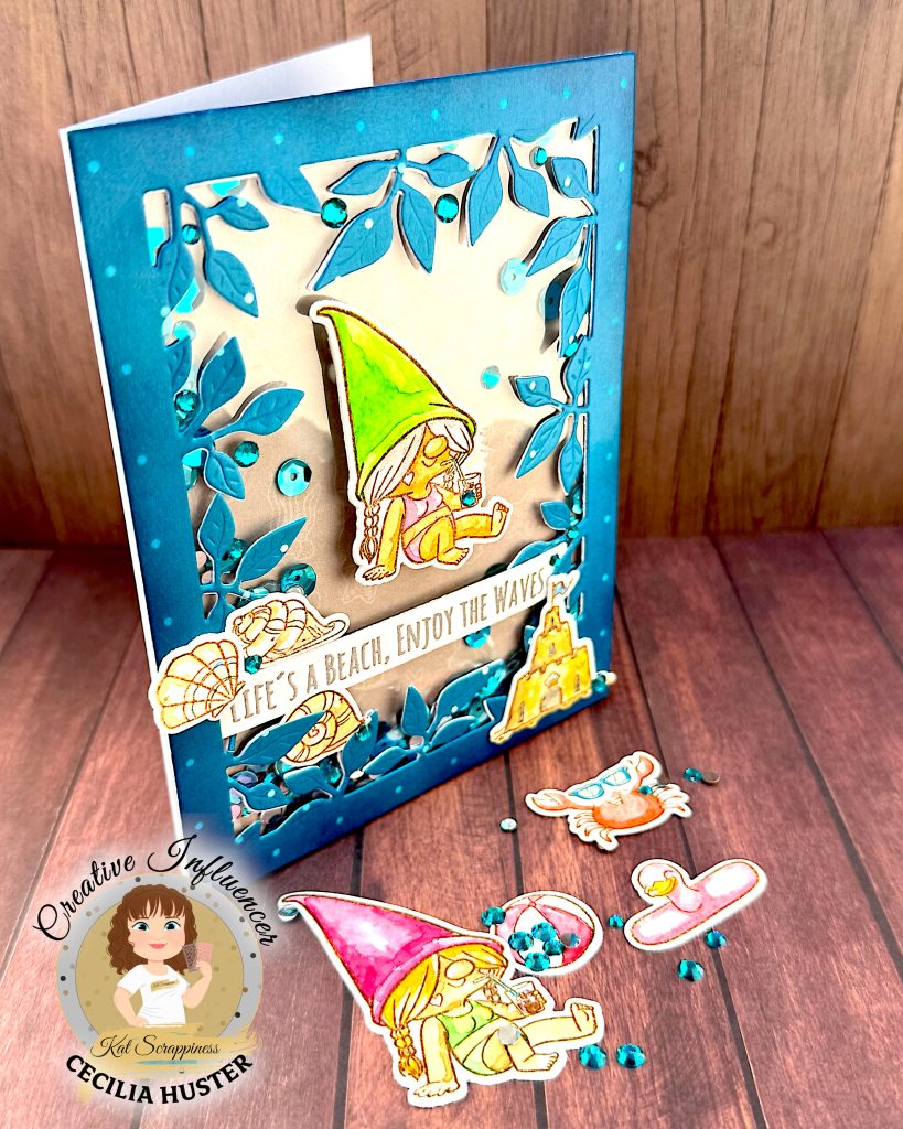 A handmade greeting card with a beach gnome theme displayed on a wooden surface. The card features a clear window filled with turquoise and silver sequins. The window is bordered by a teal, leafy frame.
The main design shows a cute gnome with long, grey hair and a bright green pointed hat. She appears to be relaxing in a bikini while holding a small drink. A banner across the card reads "LIFE'S A BEACH, ENJOY THE WAVES!" The banner is decorated with sea shells. A sandcastle has been placed in the lower right corner.
In front of the card are several coordinating die-cut elements including another gnome figure with a pink hat, a crab, and turquoise gems scattered around. The bottom left corner shows the "Kat Scrappiness Creative Influencer CECILIA HUSTER" logo featuring a cartoon woman with brown hair.