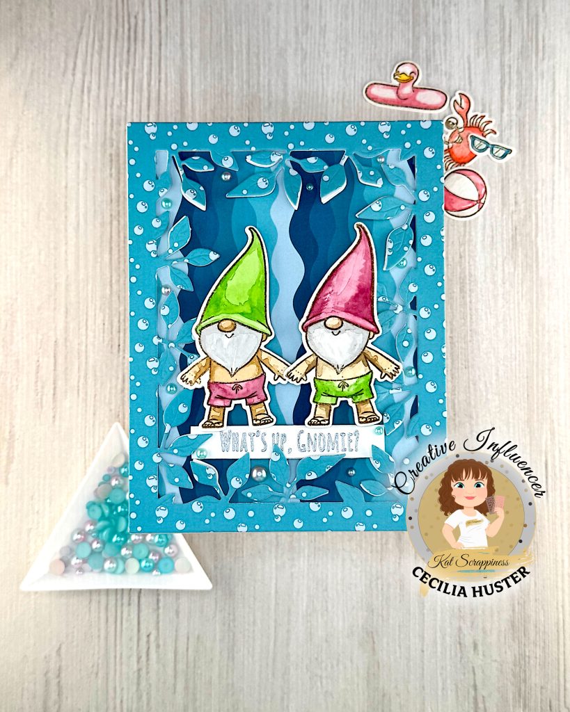 A handmade greeting card featuring two adorable gnomes with white beards wearing pointed hats - one in bright green and one in pink. The gnomes are illustrated in a cute, whimsical style and are wearing swimming trunks. They are surrounded by a leafy frame with a light blue bubble pattern.
A white banner across the bottom reads "WHAT'S UP, GNOMIES?"
In the bottom right corner is a circular logo for "Kat Scrappiness Creative Influencer CECILIA HUSTER." The card is photographed on a light wooden surface with turquoise flat backed pearls in a triangle tray in another corner. There are also some watercolored summer accessories, e.g. a pink flamingo pool float, in the background.
