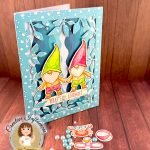 A handmade greeting card featuring two adorable gnomes with white beards wearing pointed hats - one in bright green and one in pink. The gnomes are illustrated in a cute, whimsical style and are wearing swimming trunks. They are surrounded by a leafy frame with a light blue bubble pattern. A white banner across the bottom reads "WHAT'S UP, GNOMIES?" In the bottom right corner is a circular logo for "Kat Scrappiness Creative Influencer CECILIA HUSTER." The card is standing up against a warm, brown wooden background. Some watercolored summer accessories, e.g. a pink and white beach ball, lie in the foreground. They're joined by pink and turquoise flat back pearls.
