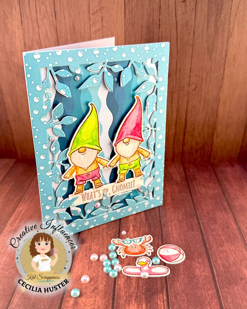 A handmade greeting card featuring two adorable gnomes with white beards wearing pointed hats - one in bright green and one in pink. The gnomes are illustrated in a cute, whimsical style and are wearing swimming trunks. They are surrounded by a leafy frame with a light blue bubble pattern.
A white banner across the bottom reads "WHAT'S UP, GNOMIES?"
In the bottom right corner is a circular logo for "Kat Scrappiness Creative Influencer CECILIA HUSTER."
The card is standing up against a warm, brown wooden background. Some watercolored summer accessories, e.g. a pink and white beach ball, lie in the foreground. They're joined by pink and turquoise flat back pearls.