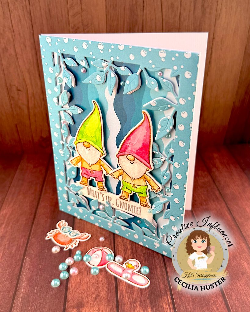 A handmade greeting card featuring two adorable gnomes with white beards wearing pointed hats - one in bright green and one in pink. The gnomes are illustrated in a cute, whimsical style and are wearing swimming trunks. They are surrounded by a leafy frame with a light blue bubble pattern.
A white banner across the bottom reads "WHAT'S UP, GNOMIES?"
In the bottom right corner is a circular logo for "Kat Scrappiness Creative Influencer CECILIA HUSTER."
The card is standing up against a warm, brown wooden background. Some watercolored summer accessories, e.g. a pink and white beach ball, lie in the foreground. They're joined by pink and turquoise flat back pearls.