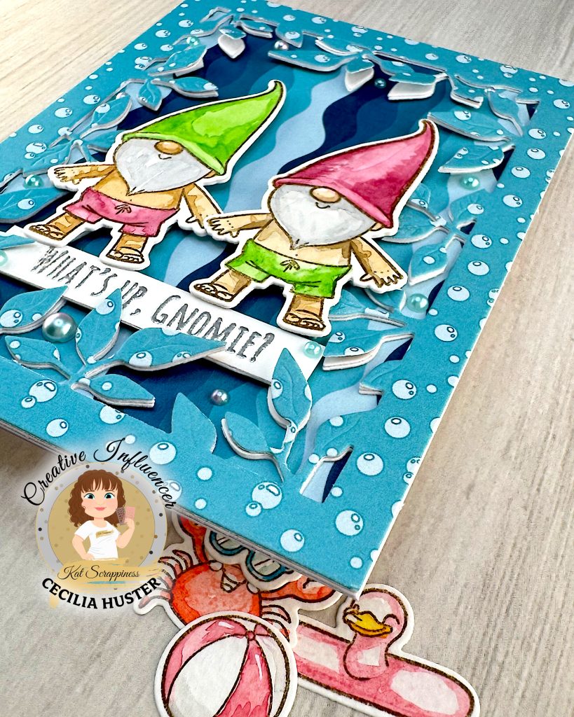 A handmade greeting card featuring two adorable gnomes with white beards wearing pointed hats - one in bright green and one in pink. The gnomes are illustrated in a cute, whimsical style and are wearing swimming trunks. They are surrounded by a leafy frame with a light blue bubble pattern.
A white banner across the bottom reads "WHAT'S UP, GNOMIES?"
In the bottom left corner is a circular logo for "Kat Scrappiness Creative Influencer CECILIA HUSTER." The card is photographed from the bottom right corner. It rests above a light wooden surface. There are some watercolored summer accessories, e.g. a pink and white beach ball, in the background.