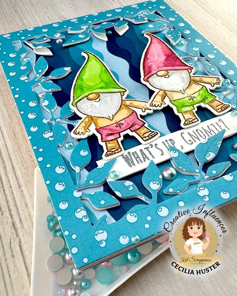 A handmade greeting card featuring two adorable gnomes with white beards wearing pointed hats - one in bright green and one in pink. The gnomes are illustrated in a cute, whimsical style and are wearing swimming trunks. They are surrounded by a leafy frame with a light blue bubble pattern.
A white banner across the bottom reads "WHAT'S UP, GNOMIES?"
In the bottom left corner is a circular logo for "Kat Scrappiness Creative Influencer CECILIA HUSTER." The card is photographed from the bottom left corner. It rests above a light wooden surface. In the foreground is a white triangle tray with turquoise and pink flat back pearls.