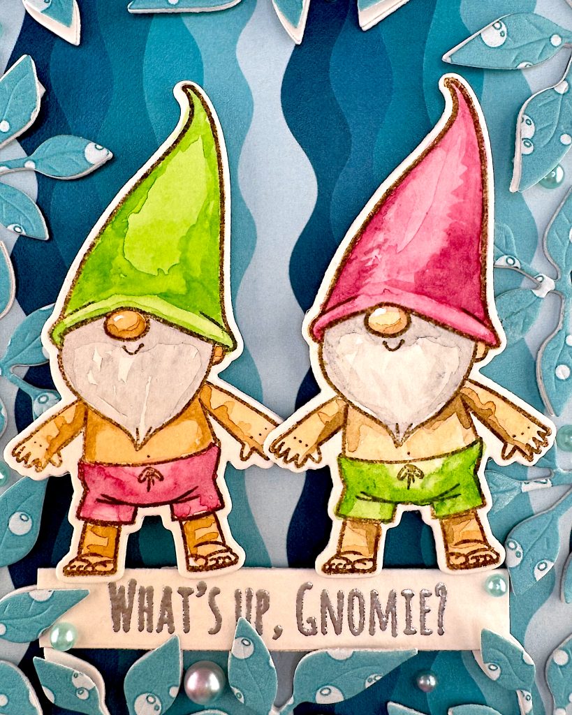 Closeup of a handmade greeting card featuring two adorable gnomes with white beards wearing pointed hats - one in bright green and one in pink. The gnomes are illustrated in a cute, whimsical style and are wearing swimming trunks.
They are surrounded by a leafy frame with a light blue bubble pattern. Turquoise and pink flat back pearls have been nestled between the leaves. The background behind them is a vertical wave pattern in blue tones.
A white banner across the bottom reads "WHAT'S UP, GNOMIES?"