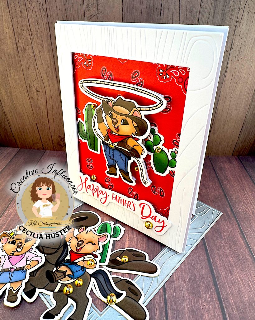 A Father's Day greeting card featuring a cute cartoon cowboy quokka character wearing a black cowboy hat, red bandana, and blue overalls. He is twirling a lasso above his head with cacti in the background. The card has a red patterned background and "Happy Father's Day" text in red script lettering.
The is standing on a wooden surface with cowboy-quokka-themed ephemera, a wood grained frame die and gold beads. The card is facing left.
There's a watermark for "Kat Scrappiness Creative Influencer CECILIA HUSTER" in the bottom left corner.
