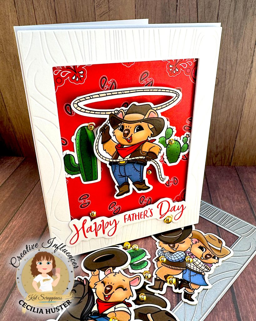 A Father's Day greeting card featuring a cute cartoon cowboy quokka character wearing a black cowboy hat, red bandana, and blue overalls. He is twirling a lasso above his head with cacti in the background. The card has a red patterned background and "Happy Father's Day" text in red script lettering.
The is standing on a wooden surface with cowboy-quokka-themed ephemera, a wood grained frame die and gold beads. The card is facing right.
There's a watermark for "Kat Scrappiness Creative Influencer CECILIA HUSTER" in the bottom left corner.