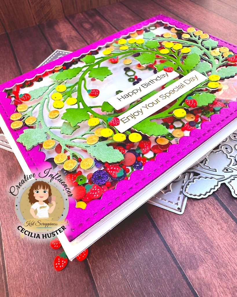 An angled view of a handmade shaker card featuring an intricate design of colorful paper embellishments arranged in a wreath pattern. The card has a bright pink scalloped border and is filled with various fruit-themed decorations including red strawberries with white dots, purple grapes, yellow citrus slices, and assorted berries in red, purple, yellow and orange.
Two white sentiment banners reading "Happy Birthday" and "Enjoy Your Special Day" in gold lettering are prominently displayed in the center. The card sits above a wooden surface with dies visible around the edges.
In the bottom left corner is a circular logo for "Creative Influencer Cecilia Huster for Kat Scrappiness."