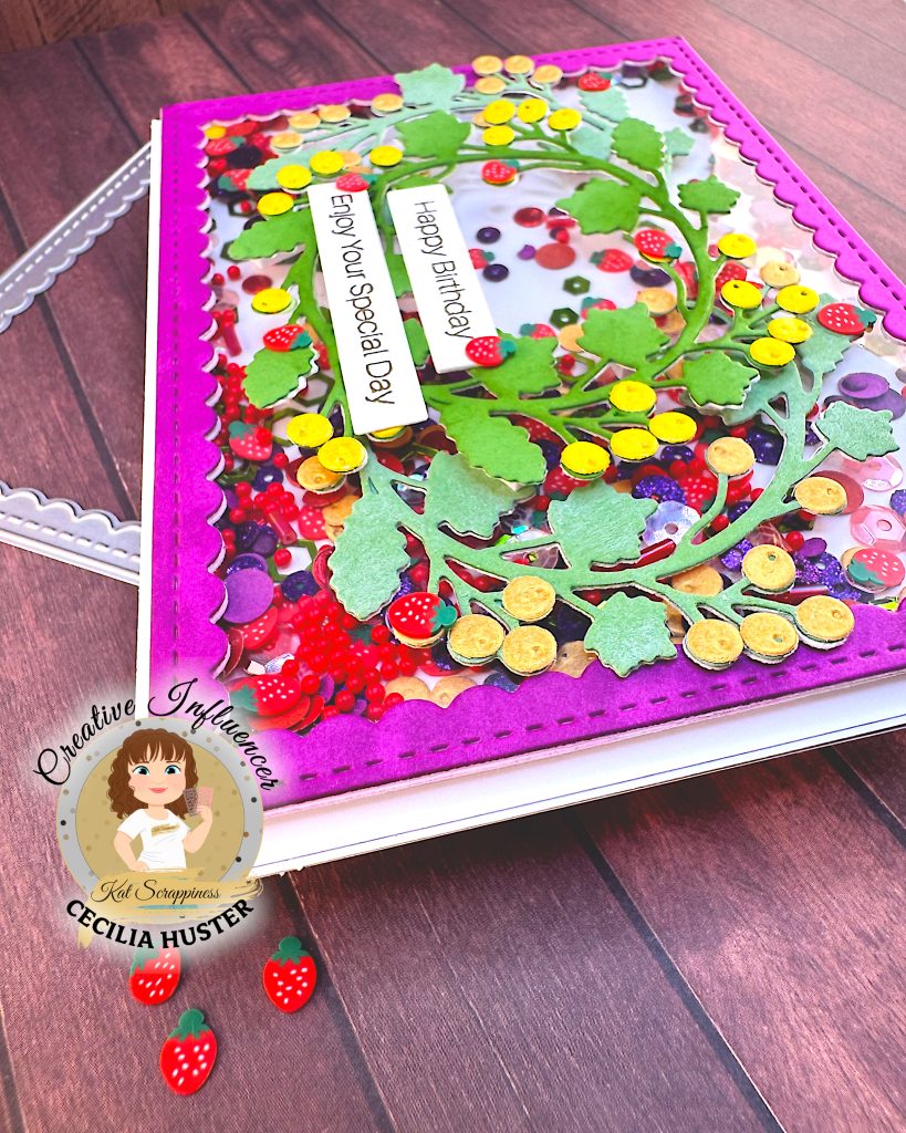 An angled view of a handmade shaker card featuring an intricate design of colorful paper embellishments arranged in a wreath pattern. The card has a bright pink scalloped border and is filled with various fruit-themed decorations including red strawberries with white dots, purple grapes, yellow citrus slices, and assorted berries in red, purple, yellow and orange.
Two white sentiment banners reading "Happy Birthday" and "Enjoy Your Special Day" in gold lettering are prominently displayed in the center. The card sits above a wooden surface with a reverse scallop frame die visible below it.
In the bottom left corner is a circular logo for "Creative Influencer Cecilia Huster for Kat Scrappiness." Below it are a few strawberry embellishments.