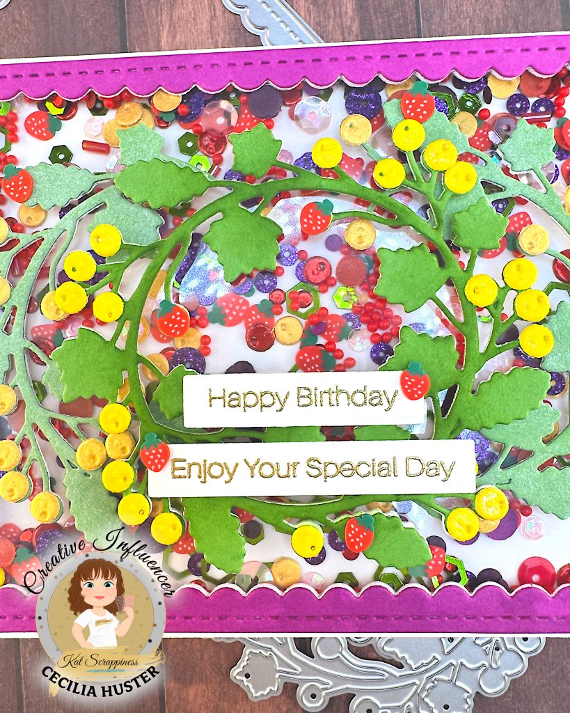 A close-up view of a handmade birthday shaker card featuring an intricate design of colorful paper embellishments arranged in a wreath pattern. The card has a bright pink scalloped border and is filled with various fruit-themed decorations including red strawberries with white dots, purple grapes, yellow citrus slices, and assorted berries in red, purple, and other colors.
Two white sentiment banners reading "Happy Birthday" and "Enjoy Your Special Day" in gold lettering are prominently displayed in the center. The card sits on a wooden surface with dies visible around the edges.
In the bottom left corner is a circular logo for "Creative Influencer Cecilia Huster for Kat Scrappiness."
