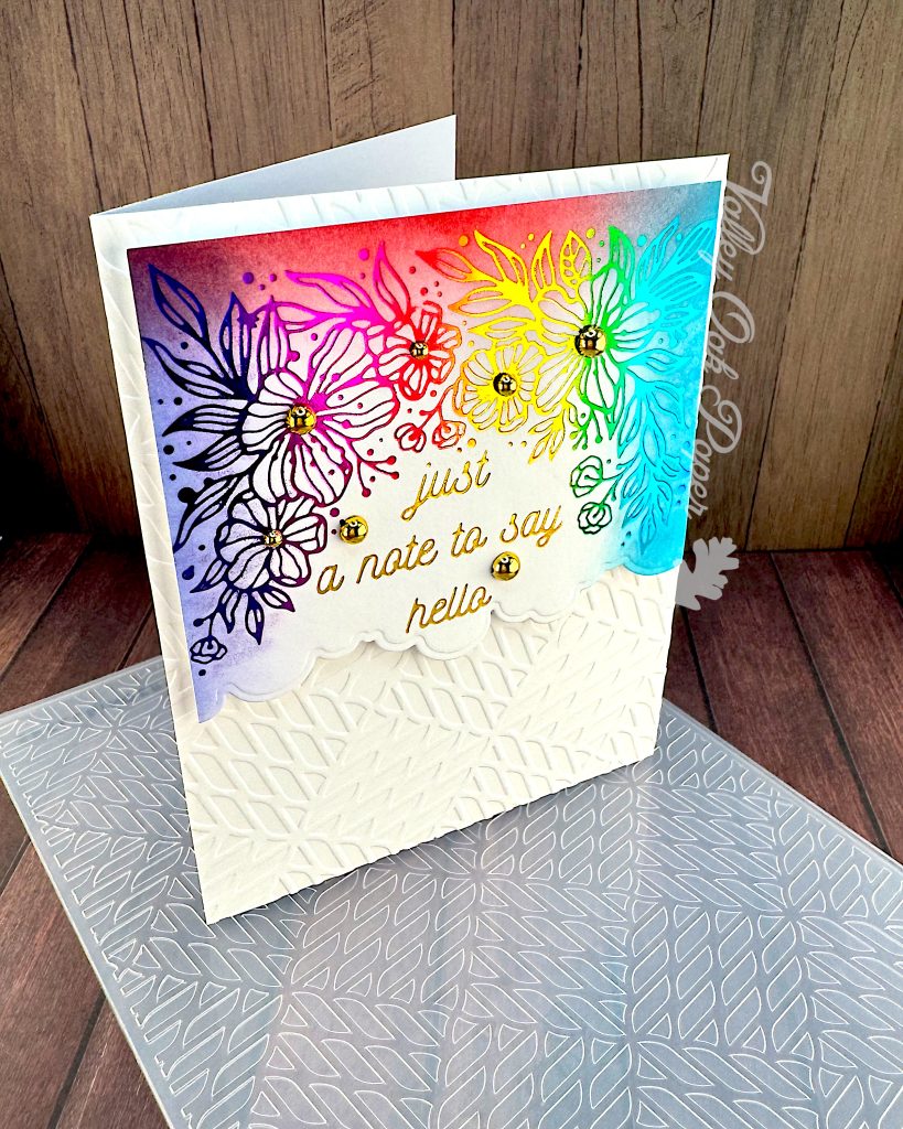 A handmade greeting card featuring a vibrant rainbow background that transitions from purple to pink, red, yellow, green, and blue. The card has a foiled line-drawn floral design overlaying the colorful background, with gold script text reading "just a note to say hello" in the center. Gold sequin accents are scattered throughout the design.
The bottom portion of the card has white embossed geometric pattern.
The card is photographed on a wooden surface alongside the embossing folder used to create the white background. There's a watermark for Valley Oak Paper visible on the right side of the image.