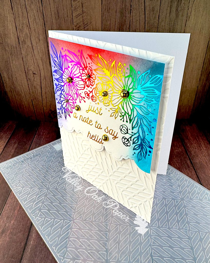 A handmade greeting card featuring a vibrant rainbow background that transitions from purple to pink, red, yellow, green, and blue. The card has a foiled line-drawn floral design overlaying the colorful background, with gold script text reading "just a note to say hello" in the center. Gold pearl accents are scattered throughout the design.
The bottom portion of the card has white embossed geometric pattern.
The card is photographed on a wooden surface alongside the embossing folder used to create the white background. There's a watermark for Valley Oak Paper visible near the bottom of the card.