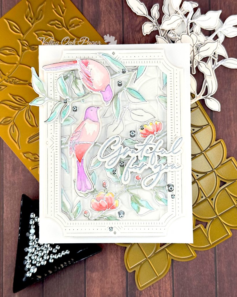 A handmade greeting card featuring watercolor illustrations of pink and purple birds among delicate green foliage and coral-colored flowers.
The card has an ornate white decorative border with small gem accents, and includes cursive text that says "Grateful for You."
The card is photographed on a wooden surface alongside crafting supplies including golden hot foiling plates with leaf patterns, white die-cut decorative elements, and a black triangle tray containing embellishments.
