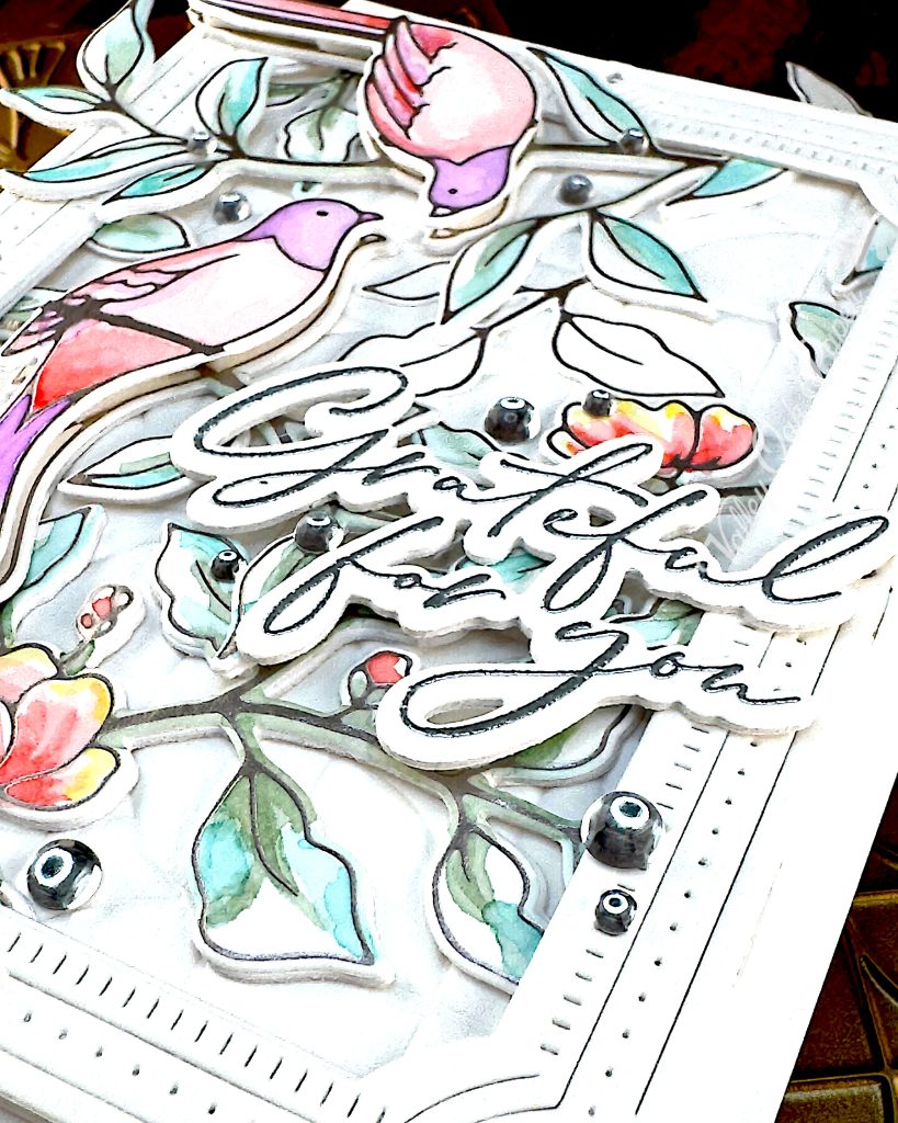 A handmade greeting card featuring watercolor illustrations of pink and purple birds among delicate green foliage and coral-colored flowers.
The card has an ornate white decorative border with small gem accents, and includes cursive text that says "Grateful for You."
This angle highlights the sentiment and the stunning dimension of the card.