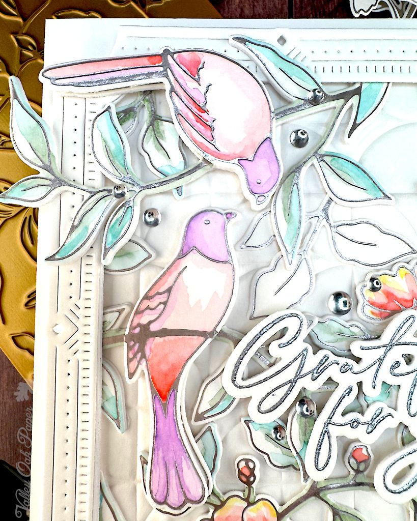 A handmade greeting card featuring watercolor illustrations of pink and purple birds among delicate green foliage and coral-colored flowers.
The card has an ornate white decorative border with small gem accents, and includes cursive text that says "Grateful for You."
This closeup shows the watercolored birds in detail. They appear to be interacting with each other. The topmost bird is bending down toward the other bird.