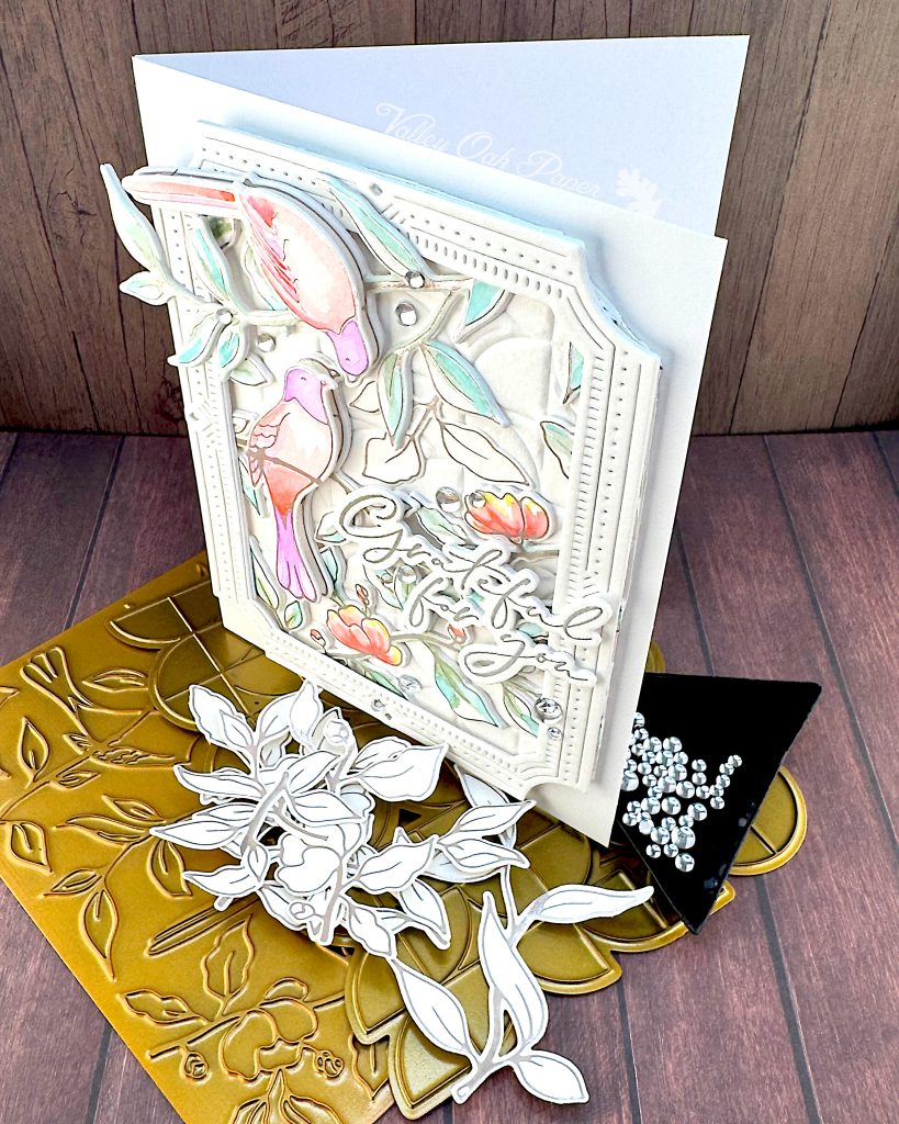 A handmade greeting card featuring watercolor illustrations of pink and purple birds among delicate green foliage and coral-colored flowers.
The card has an ornate white decorative border with small gem accents, and includes cursive text that says "Grateful for You."
The card is standing facing left on a golden hot foiling plate used to create it. In front of it are some leftover white die cuts with silver hot foiling. Behind the card is a black triangle tray containing clear dew drops.