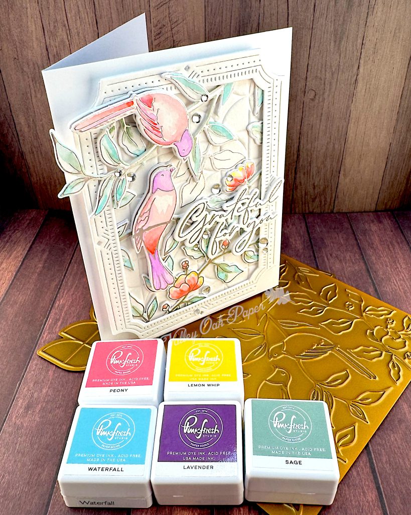 A handmade greeting card featuring watercolor illustrations of pink and purple birds among delicate green foliage and coral-colored flowers.
The card has an ornate white decorative border with small gem accents, and includes cursive text that says "Grateful for You."
The card is standing on a golden hot foiling plate used to create it. In front of it are five Pinkfresh Studio Mini Ink Pads in the colors Peony, Lemon Whip, Waterfall, Lavender and Sage.