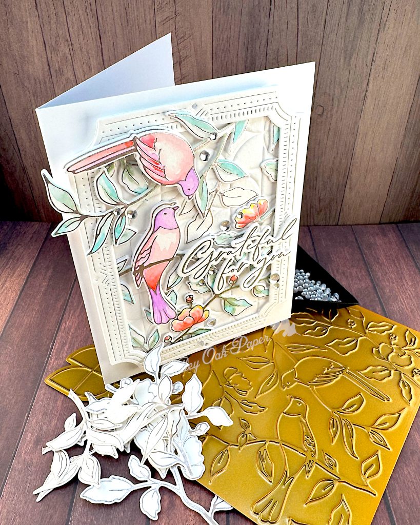 A handmade greeting card featuring watercolor illustrations of pink and purple birds among delicate green foliage and coral-colored flowers.
The card has an ornate white decorative border with small gem accents, and includes cursive text that says "Grateful for You."
The card is standing facing right on a golden hot foiling plate used to create it. In front of it are some leftover white die cuts with silver hot foiling. Behind the card is a black triangle tray containing clear dew drops.