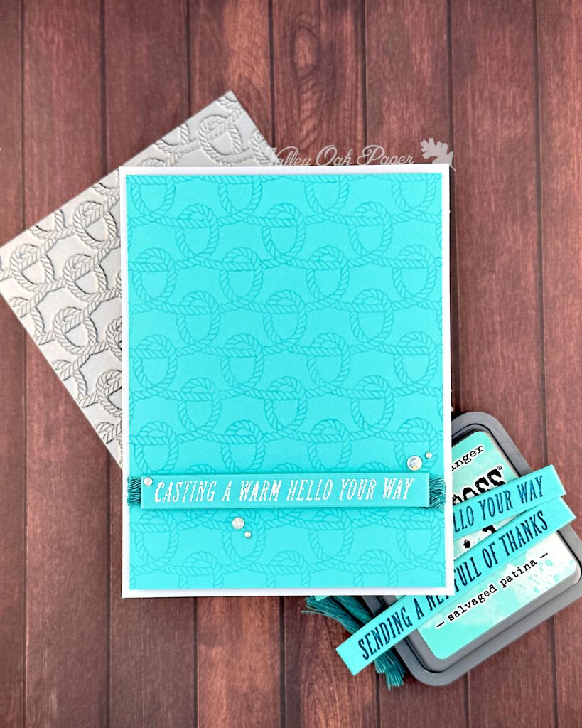 A handmade greeting card with a bright turquoise background featuring an embossed rope knot pattern.
The card has white text reading "CASTING A WARM HELLO YOUR WAY" on a banner across the bottom, with small white pearl embellishments.
Behind the card is the press plate used to create the rope knot pattern. To the right is an ink pad in the color Salvaged Patina with turquoise sentiment strips visible.
The scene is photographed on a wooden surface with a "Valley Oak Paper" watermark visible in the top right corner.