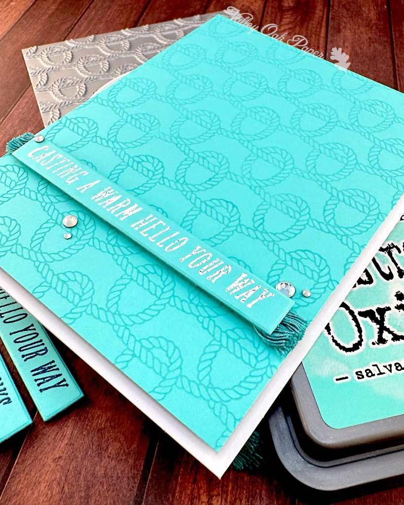 A handmade greeting card with a bright turquoise background featuring an embossed rope knot pattern seen from the bottom right corner.
The card has white text reading "CASTING A WARM HELLO YOUR WAY" on a banner across the bottom, with small white gem embellishments.
Under the card is the press plate used to create the rope knot pattern. To the right is a Distress Oxide ink pad in the color Salvaged Patina. Some sentiment strips are visible to the left of the card.
The scene is photographed on a wooden surface with a "Valley Oak Paper" watermark visible in the top right corner.