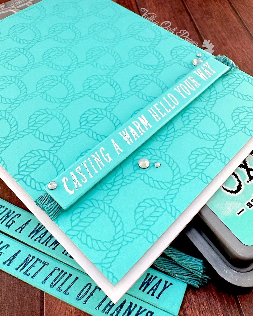 A handmade greeting card with a bright turquoise background featuring an embossed rope knot pattern seen from the bottom left corner.
The card has white text reading "CASTING A WARM HELLO YOUR WAY" on a banner across the bottom, with small white gem embellishments.
To the right is a Distress Oxide ink pad in the color Salvaged Patina. Some sentiment strips and teal macramé cords are visible under the card.
The scene is photographed on a wooden surface with a "Valley Oak Paper" watermark visible in the top right corner.