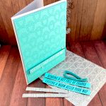 A handmade greeting card with a bright turquoise background featuring an embossed rope knot pattern is standing, facing right, on a wooden surface. The card has white text reading "CASTING A WARM HELLO YOUR WAY" on a banner across the bottom, with small white pearl embellishments around it. The card stands on the press plate used to create the rope knot pattern. Some turquoise sentiment strips with the texts "Sending a Net Full of Thanks" and "Casting a Warm Hello Your Way" lie on the press plate. Above them are two ends of turquoise macramé cordage. It's the same cordage as used behind the sentiment on the card. The two small press plates with the sentiments are also present. The scene is photographed on a wooden surface with a "Valley Oak Paper" watermark visible in the top right corner.