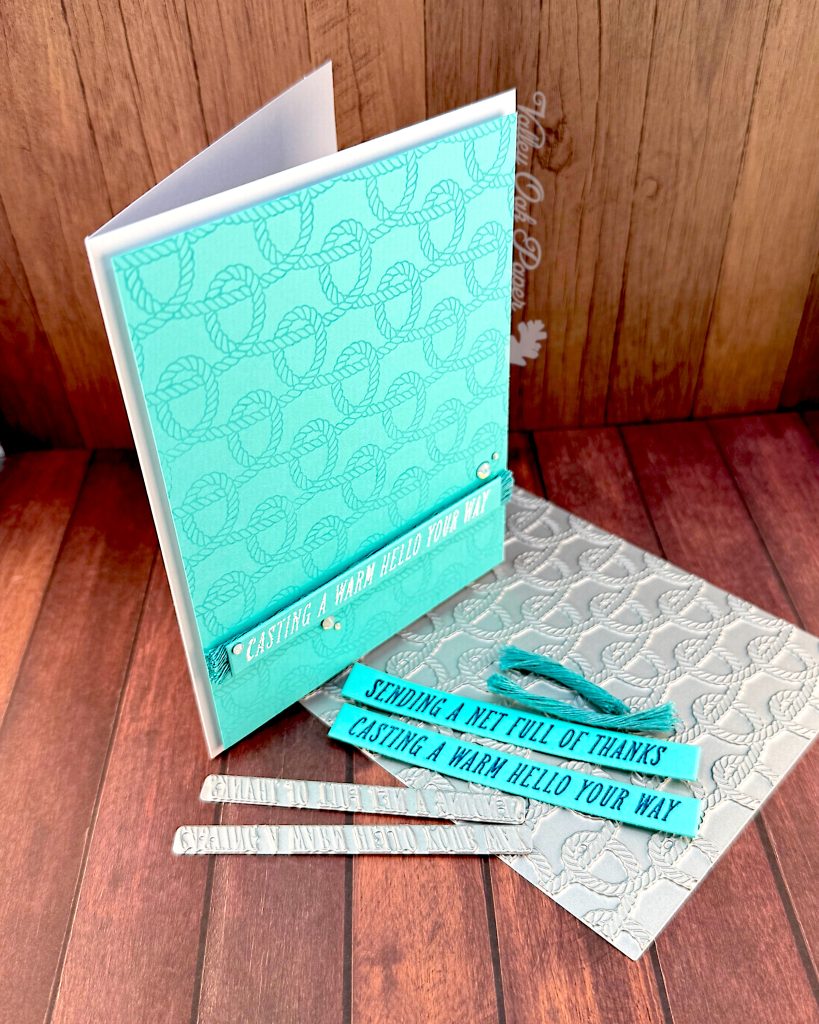 A handmade greeting card with a bright turquoise background featuring an embossed rope knot pattern is standing, facing right, on a wooden surface.
The card has white text reading "CASTING A WARM HELLO YOUR WAY" on a banner across the bottom, with small white pearl embellishments around it.
The card stands on the press plate used to create the rope knot pattern. Some turquoise sentiment strips with the texts "Sending a Net Full of Thanks" and "Casting a Warm Hello Your Way" lie on the press plate. Above them are two ends of turquoise macramé cordage. It's the same cordage as used behind the sentiment on the card.
The two small press plates with the sentiments are also present.
The scene is photographed on a wooden surface with a "Valley Oak Paper" watermark visible in the top right corner.