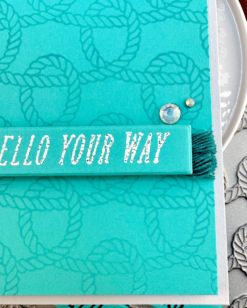A detail of a handmade greeting card with a bright turquoise background featuring an embossed rope knot pattern. Only the bottom right corner is visible.
The card has a sentiment with white text across the bottom. The banner rests on turquoise cordage. Two small white gem embellishments are visible above the banner.
Under the card can be glimpsed the press plate used to create the rope knot pattern.
A "Valley Oak Paper" watermark visible in the top right corner.
