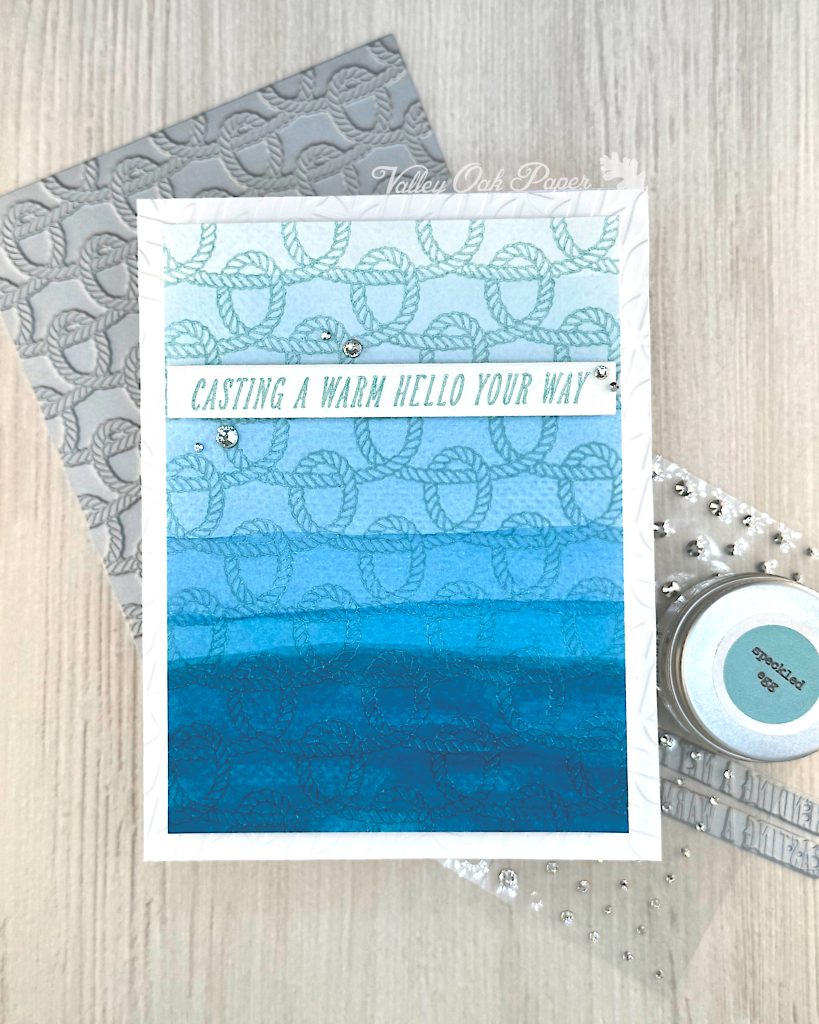 A handmade greeting card with a nautical theme featuring a faux dip dye blue gradient background that transitions from light blue at the top to deep ocean blue at the bottom. 
The card is decorated with an embossed rope pattern throughout, creating texture and visual interest. A white banner across the middle contains the message "CASTING A WARM HELLO YOUR WAY" in turquoise lettering. 

The card is embellished with small silver rhinestones scattered across the surface. Behind the main card, there's a gray BetterPress plate with the same rope pattern as on the card. The scene is photographed on a light wood surface with crafting supplies visible, including a jar of Speckled Egg Distress Embossing Glaze and additional rhinestone embellishments.