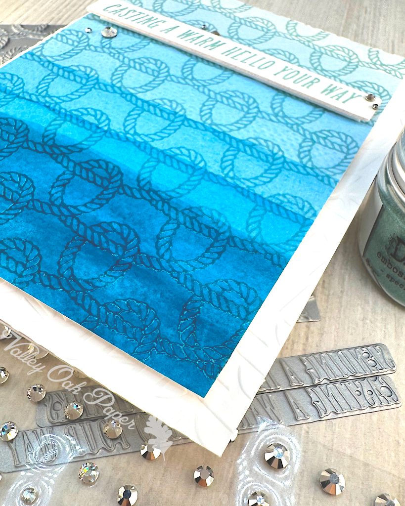 A handmade greeting card with a nautical theme is seen from the lower right corner. The card features a faux dip dye blue gradient background that transitions from light blue at the top to deep ocean blue at the bottom. 

It's decorated with an embossed rope pattern throughout, creating texture and visual interest. A white banner across the middle contains the message "CASTING A WARM HELLO YOUR WAY" in turquoise lettering. 

The card is embellished with small silver rhinestones scattered across the surface. Under the card, there are BetterPress sentiment plates and additional rhinestone embellishments. The scene is photographed on a light wood surface.