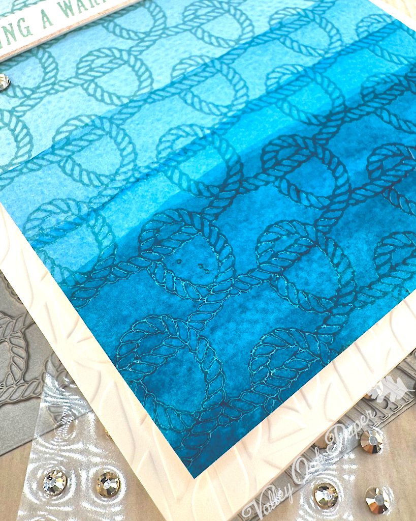A handmade greeting card with a nautical theme is seen from the lower left corner. The card features a faux dip dye blue gradient background that transitions from light blue at the top to deep ocean blue at the bottom. 

It's decorated with an embossed rope pattern throughout, creating texture and visual interest. 

Under the card, there are BetterPress plates and additional rhinestone embellishments. The scene is photographed on a light wood surface.