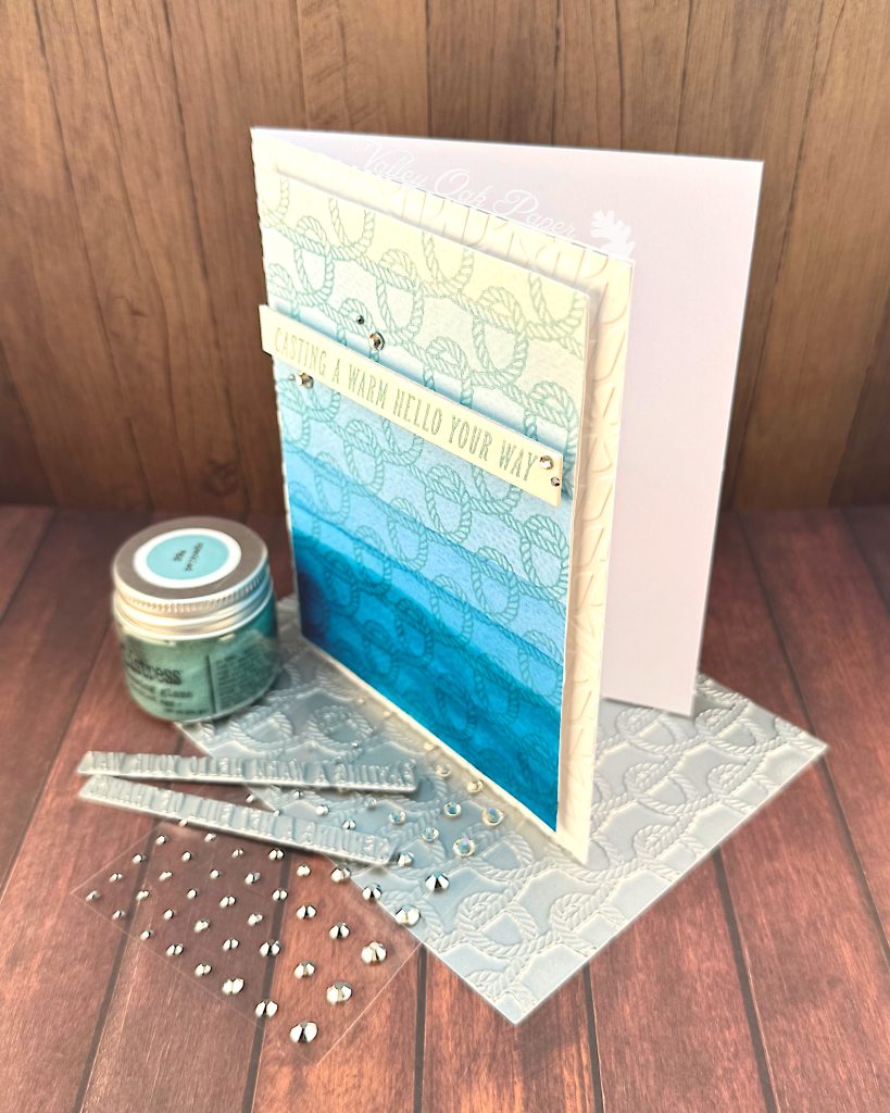 A handmade greeting card with a nautical theme featuring a faux dip dye blue gradient background that transitions from light blue at the top to deep ocean blue at the bottom. 
The card is decorated with an embossed rope pattern throughout, creating texture and visual interest. A white banner across the middle contains the message "CASTING A WARM HELLO YOUR WAY" in turquoise lettering. 

The card is embellished with small silver rhinestones scattered across the surface. It's standing on a BetterPress plate with the same rope pattern as on the card. The scene is photographed on a against dark wood surfaces with crafting supplies visible, including a jar of Speckled Egg Distress Embossing Glaze and additional rhinestone embellishments.