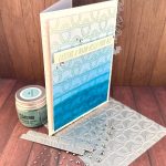 A handmade greeting card with a nautical theme featuring a faux dip dye blue gradient background that transitions from light blue at the top to deep ocean blue at the bottom. The card is decorated with an embossed rope pattern throughout, creating texture and visual interest. A white banner across the middle contains the message "CASTING A WARM HELLO YOUR WAY" in turquoise lettering. The card is embellished with small silver rhinestones scattered across the surface. It's standing on a BetterPress plate with the same rope pattern as on the card. The scene is photographed on a against dark wood surfaces with crafting supplies visible, including a jar of Speckled Egg Distress Embossing Glaze and additional rhinestone embellishments.