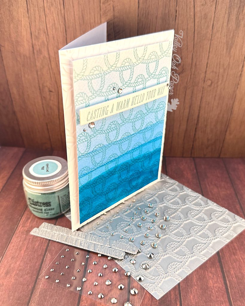 A handmade greeting card with a nautical theme featuring a faux dip dye blue gradient background that transitions from light blue at the top to deep ocean blue at the bottom. 
The card is decorated with an embossed rope pattern throughout, creating texture and visual interest. A white banner across the middle contains the message "CASTING A WARM HELLO YOUR WAY" in turquoise lettering. 

The card is embellished with small silver rhinestones scattered across the surface. It's standing on a BetterPress plate with the same rope pattern as on the card. The scene is photographed on a against dark wood surfaces with crafting supplies visible, including a jar of Speckled Egg Distress Embossing Glaze and additional rhinestone embellishments.