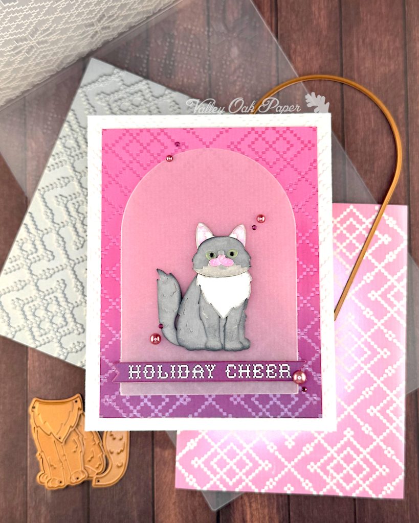 A handmade holiday greeting card featuring a gray and white cat die cut sitting on a vellum arch. The card has a pink and purple ombre background with a pink crosshatch pattern and "HOLIDAY CHEER" in white text at the bottom. The card is white and textured.
It's displayed on a wooden surface alongside the BetterPress plate that created the crosshatch pattern, the embossing folder that created the texture, an arch die and some of the dies used to create the cat. The "Valley Oak Paper" watermark is visible in a corner.