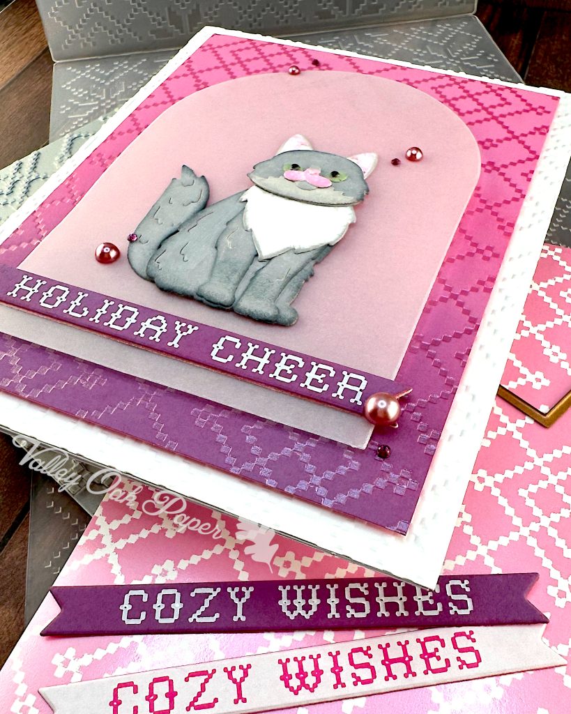 A handmade holiday greeting card featuring a gray and white cat die cut sitting on a vellum arch. The card has a pink and purple ombre background with a pink crosshatch pattern and "HOLIDAY CHEER" in white text at the bottom. The card is white and textured. It has been photographed from the bottom right corner.
It's displayed on a wooden surface alongside the BetterPress plate that created the crosshatch pattern, the embossing folder that created the texture, an arch die and two sentiment banners with the text "Cozy Wishes." The Valley Oak Paper watermark is visible in a corner.