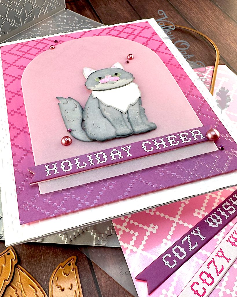 A handmade holiday greeting card featuring a gray and white cat die cut sitting on a vellum arch. The card has a pink and purple ombre background with a pink crosshatch pattern and "HOLIDAY CHEER" in white text at the bottom. The card is white and textured. It has been photographed from the bottom left corner.
It's displayed on a wooden surface alongside the BetterPress plate that created the crosshatch pattern, the embossing folder that created the texture, an arch die, some cat dies and two sentiment banners with the text "Cozy Wishes." The Valley Oak Paper watermark is visible in a corner.