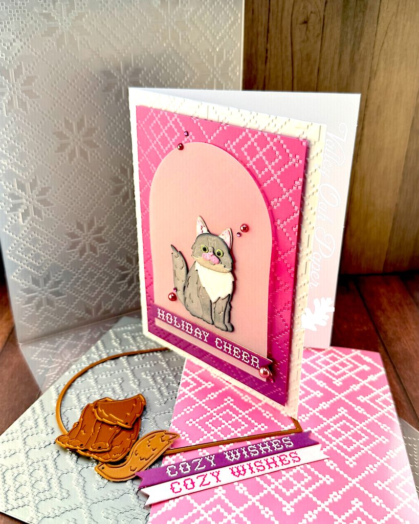 A handmade holiday greeting card stands facing left. It features a gray and white cat die cut sitting on a vellum arch. The card has a pink and purple ombre background with a pink crosshatch pattern and "HOLIDAY CHEER" in white text at the bottom. The card is white and textured.
It's displayed on a wooden surface alongside the BetterPress plate that created the crosshatch pattern, the embossing folder that created the texture, an arch die, some cat dies and two sentiment banners with the text "Cozy Wishes." The Valley Oak Paper watermark is visible in a corner.