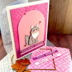 A handmade holiday greeting card featuring a gray and white cat die cut sitting on a vellum arch. The card has a pink and purple ombre background with a pink crosshatch pattern and "HOLIDAY CHEER" in white text at the bottom. The card is white and textured. It has been photographed from the bottom right corner. It's displayed on a wooden surface alongside the BetterPress plate that created the crosshatch pattern, the embossing folder that created the texture, an arch die and two sentiment banners with the text "Cozy Wishes." The Valley Oak Paper watermark is visible in a corner.