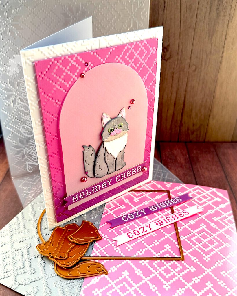 A handmade holiday greeting card stands facing right. It features a gray and white cat die cut sitting on a vellum arch. The card has a pink and purple ombre background with a pink crosshatch pattern and "HOLIDAY CHEER" in white text at the bottom. The card is white and textured.
It's displayed on a wooden surface alongside the BetterPress plate that created the crosshatch pattern, the embossing folder that created the texture, an arch die, some cat dies and two sentiment banners with the text "Cozy Wishes." The Valley Oak Paper watermark is visible in a corner.