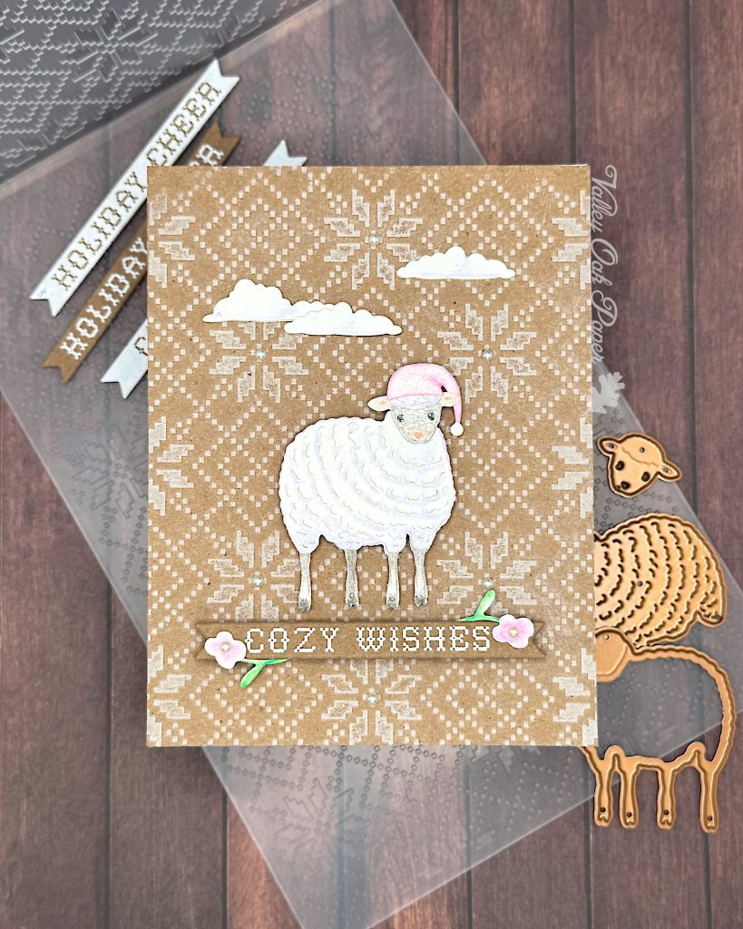 A handmade holiday greeting card featuring a cute white sheep wearing a pink Santa hat on a Kraft background with an embossed Nordic knit pattern. The card includes white die-cut clouds in the sky and a banner reading "COZY WISHES" decorated with small floral elements. The scene is accented with small pearls. The card is floating above a wooden surface.

Between the card and the wooden surface, there are white paper banner strips with text including "COZY WISHES" and "HOLIDAY CHEER," along with metal dies showing a sheep outline and a sheep silhouette. An embossing folder with a matching Nordic knit pattern can also be seen. 