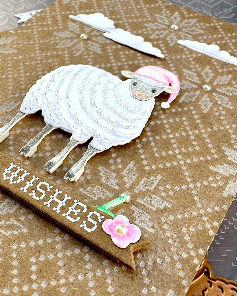 A handmade holiday greeting card featuring a cute white sheep wearing a pink Santa hat on a Kraft background with an embossed Nordic knit pattern. The card includes white die-cut clouds in the sky and a banner  decorated with small floral elements. The scene is accented with small pearls. The card is lying on the embossing folder that created the Nordic knit pattern. 
