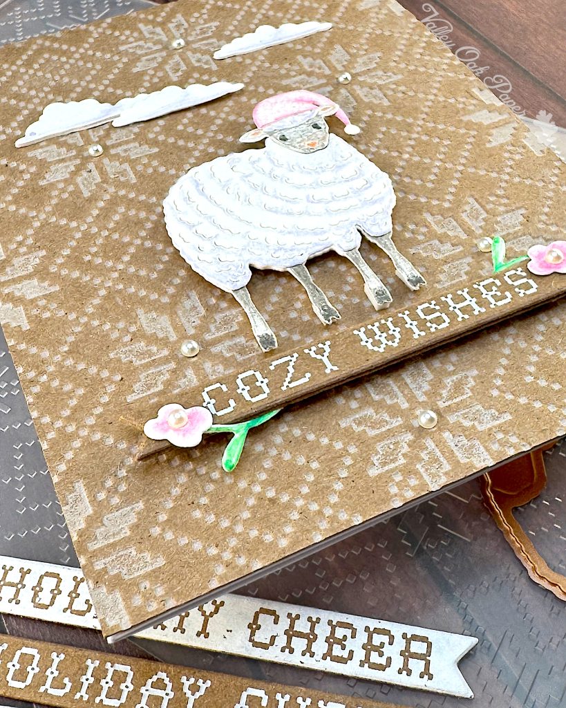 A handmade holiday greeting card featuring a cute white sheep wearing a pink Santa hat on a Kraft background with an embossed Nordic knit pattern. The card includes white die-cut clouds in the sky and a banner reading "COZY WISHES" decorated with small floral elements. The scene is accented with small pearls. The card is lying on the embossing folder that created the Nordic knit pattern. 

In the foreground, in front of the card, there are white paper banner strips with text including  "HOLIDAY CHEER."
