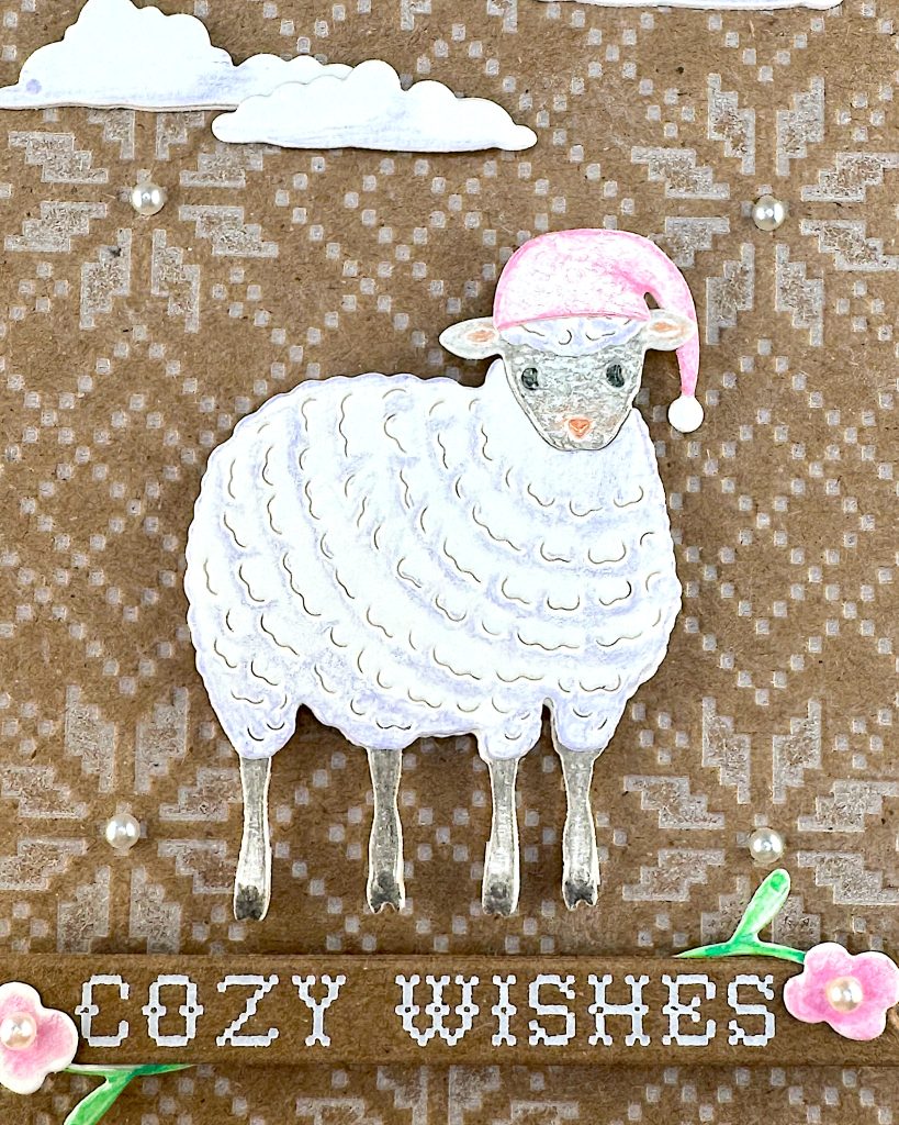 A closeup of a handmade holiday greeting card. It features a cute white sheep wearing a pink Santa hat on a Kraft background with an embossed Nordic knit pattern. The card includes white die-cut clouds in the sky and a banner reading "COZY WISHES" decorated with small floral elements. The scene is accented with small pearls. 