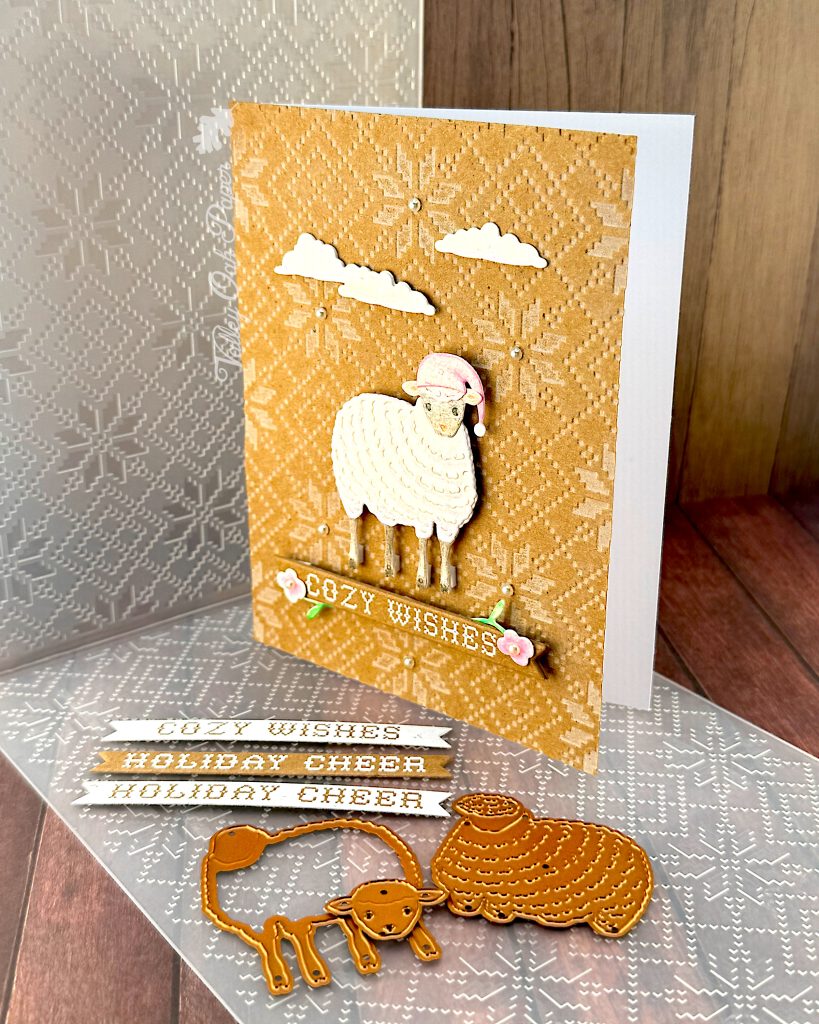A handmade holiday greeting card featuring a cute white sheep wearing a pink Santa hat on a Kraft background with an embossed Nordic knit pattern. The card includes white die-cut clouds in the sky and a banner reading "COZY WISHES" decorated with small floral elements. The scene is accented with small pearls. The card is standing up facing left. 

In the foreground, in front of the card, there are white paper banner strips with text including "COZY WISHES" and "HOLIDAY CHEER," along with metal dies showing a sheep outline and a sheep silhouette. Behind the card is an embossing folder with a matching Nordic knit pattern. The entire scene is photographed on a wooden surface.