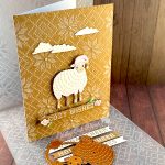 A handmade holiday greeting card featuring a cute white sheep wearing a pink Santa hat on a Kraft background with an embossed Nordic knit pattern. The card includes white die-cut clouds in the sky and a banner reading "COZY WISHES" decorated with small floral elements. The scene is accented with small pearls. The card is standing up facing right. In the foreground, in front of the card, there are white paper banner strips with text including "COZY WISHES" and "HOLIDAY CHEER," along with metal dies showing a sheep outline and a sheep silhouette. Behind the card is an embossing folder with a matching Nordic knit pattern. The entire scene is photographed on a wooden surface.