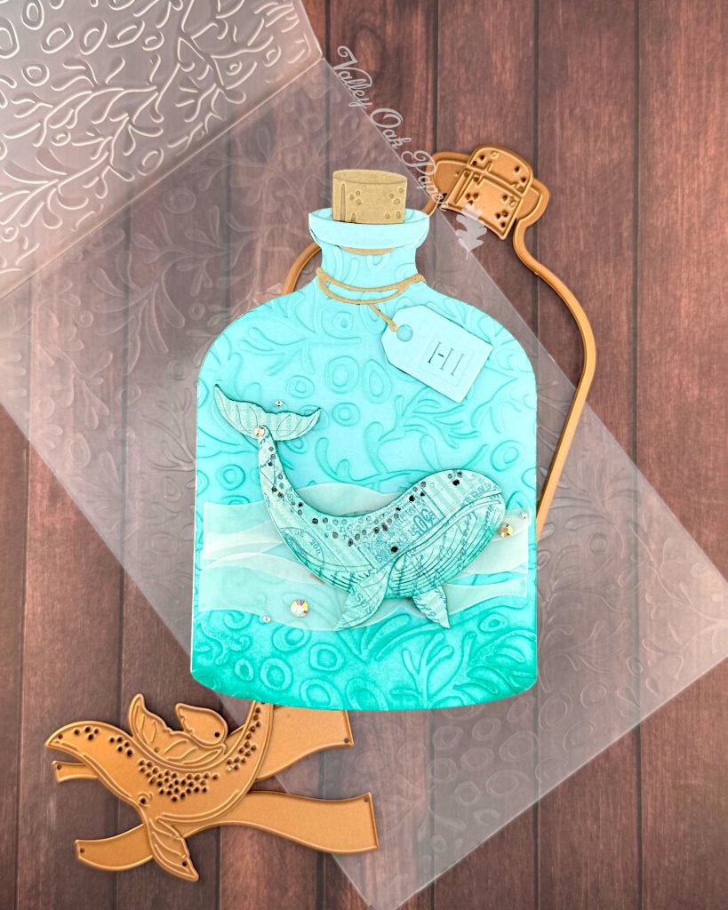 A handmade greeting card in the shape of a turquoise glass bottle with a cork stopper and "Hi" tag. The bottle contains a humpback whale swimming in stylized vellum ocean waves. The bottle has been embossed with a coral pattern.
It is surrounded by some of the dies sea used when creating it. The card sits on the coral pattern embossing folder used to create it. It is pictured on a wooden surface. In the top center, the Valley Oak Paper logo is visible.